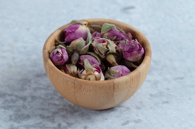 Boutons de roses séchées rouge rose dans un bol en bois avec des pétales placés sur une pierre.