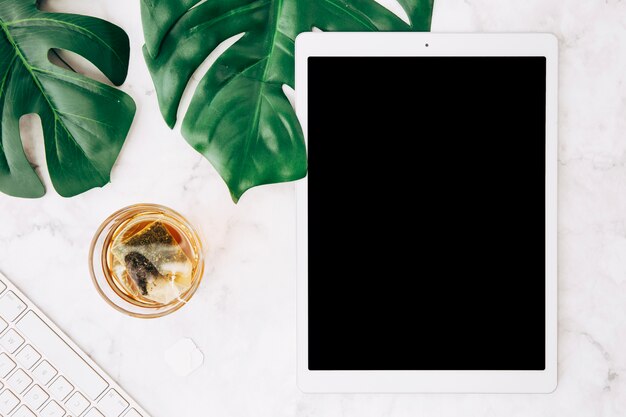 Brassage de boisson chaude avec un sachet de thé en verre; feuilles de monstera; clavier et tablette numérique sur bureau blanc