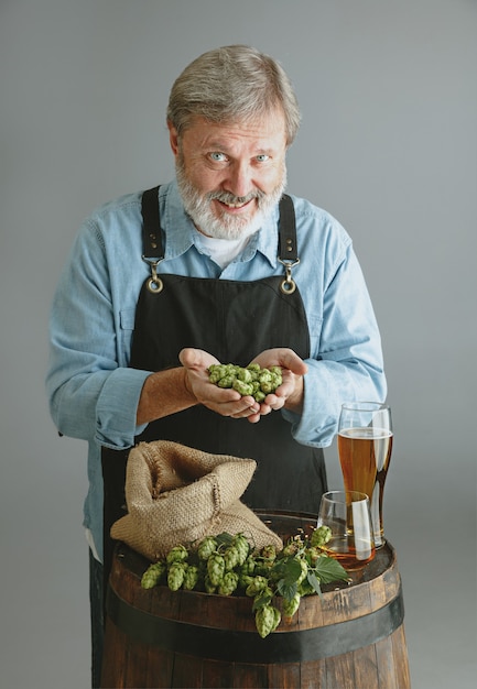 Photo gratuite brasseur d'homme senior confiant avec bière artisanale en verre sur tonneau en bois sur mur gris. le propriétaire de l'usine a présenté ses produits, testant la qualité.