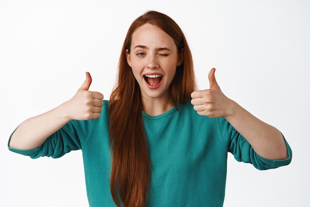 Photo gratuite bravo une rousse souriante qui fait un clin d'œil et montre les pouces vers le haut comme et approuve les éloges beau travail excellent geste de travail faisant allusion à quelque chose de bon recommander le service