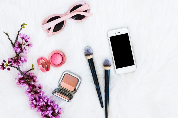 Brindille de fleur pourpre avec une poudre faciale compacte; pinceaux à maquillage; téléphone portable et lunettes de soleil sur fond de fourrure