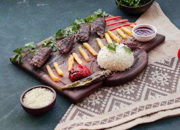 Brochette de boeuf, bâtonnets de pomme de terre frits, grillades, garniture de riz et sauce sur une planche de bois.