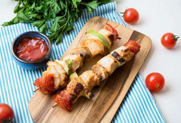 Brochettes de poulet à angle élevé sur une planche à découper avec des sauces et des tomates