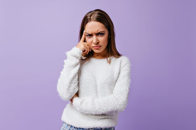 Brune émotionnelle avec colère et indignation pose pour portrait féminin sur lilas. Malheureuse fille en pull en laine touche son visage