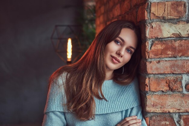 Une brune sensuelle dans un pull gris appuyée contre un mur de briques dans une pièce avec un intérieur loft.