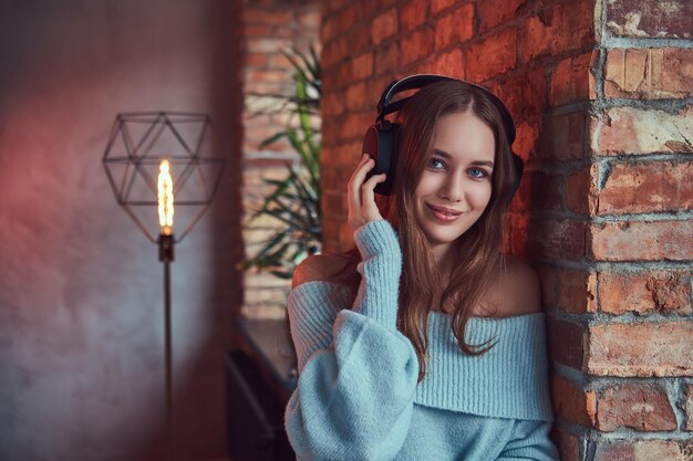 Une brune sensuelle dans un pull gris avec un casque appuyé contre un mur de briques dans une pièce avec un intérieur loft.