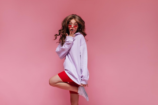 Brunette bouclée femme en rouge lunettes de soleil violet sweat à capuche et short de cyclisme saute sur isolé Cool girl semble surpris sur fond rose