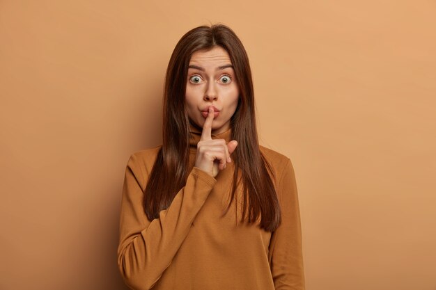 Brunette femme européenne garde l'index sur les lèvres, fait un geste silencieux, garde le secret, choqué par les rumeurs, a une peau saine, porte un pull marron décontracté