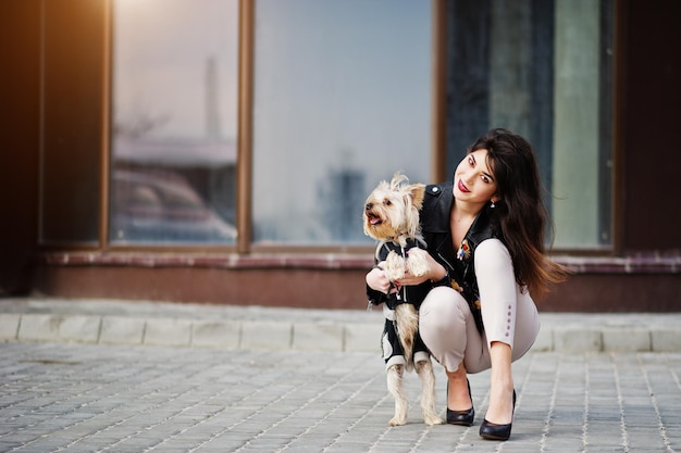Photo gratuite brunette gitane avec un chien yorkshire terrier posé contre de grandes fenêtres maison modèle porter sur une veste en cuir et un t-shirt avec un pantalon d'ornement et des chaussures à talons hauts