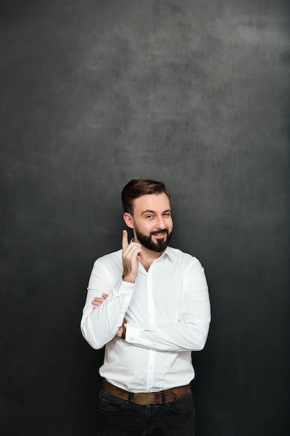 Brunette homme d'affaires posant devant la caméra avec un regard heureux délicat, pointant l'index comme s'il savait quelque chose sur gris foncé