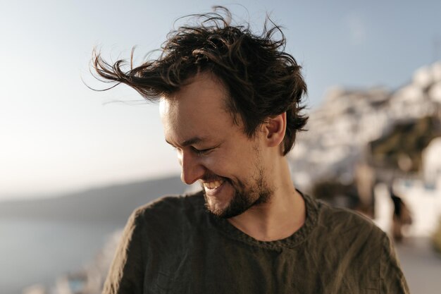 Brunette homme en t-shirt vert foncé sourit largement à l'extérieur Closeup portrait of young guy rit sur fond de mer