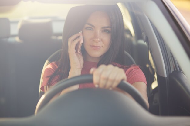 Brunette jeune conductrice parle via un téléphone portable moderne en conduisant une voiture, a une grave expression