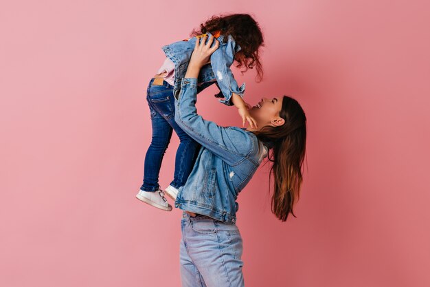 Brunette jeune femme tenant sa fille sur fond rose. Photo de Studio de maman et enfant préadolescent dans des vestes en jean.
