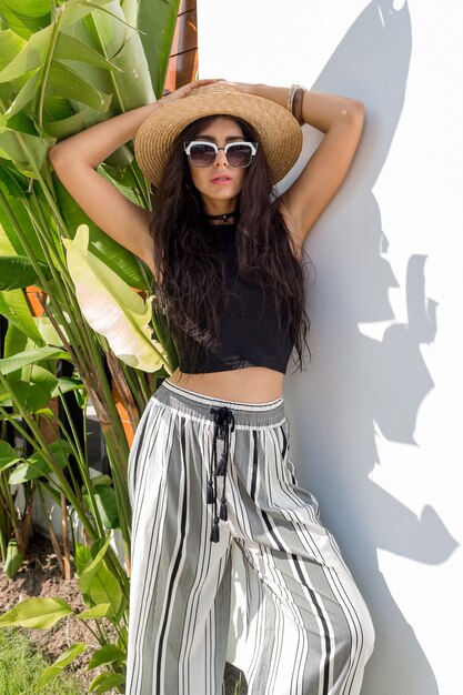 Brunette sexy avec un corps bronzé en chapeau de paille posant en plein air. Tendances de la mode estivale. Ambiance tropicale. Bracelets en bois.