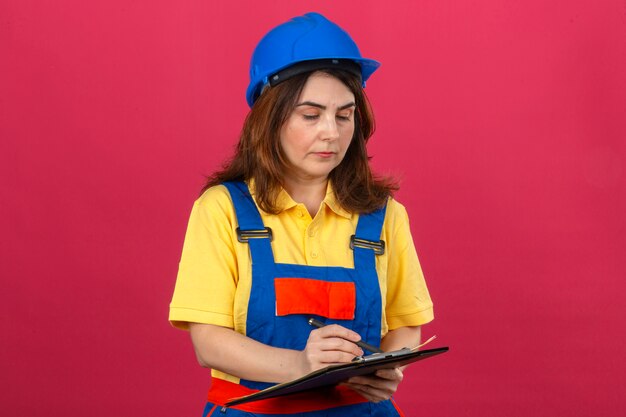 Builder femme portant des uniformes de construction et un casque de sécurité tenant le presse-papiers en le regardant avec un visage sérieux sur un mur rose isolé