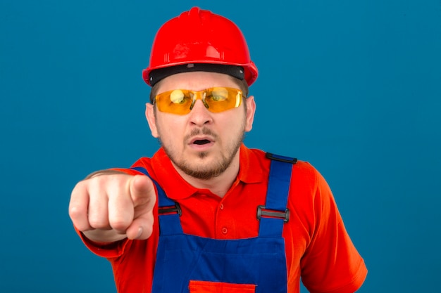 Builder homme portant l'uniforme de construction et casque de sécurité choqué pointant avec le doigt à la caméra sur mur bleu isolé