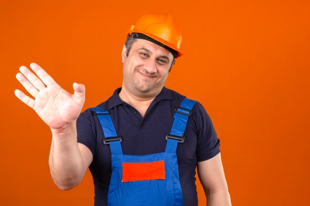 Builder homme portant un uniforme de construction et un casque de sécurité souriant et heureux en agitant une main sur un mur orange isolé