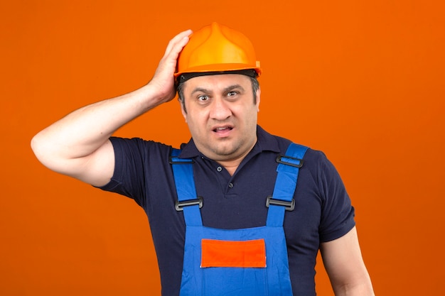 Builder Homme Portant Des Uniformes De Construction Et Un Casque De Sécurité à La Confusion Et Déçu De N'avoir Aucune Idée De Toucher Son Casque Sur Un Mur Orange Isolé