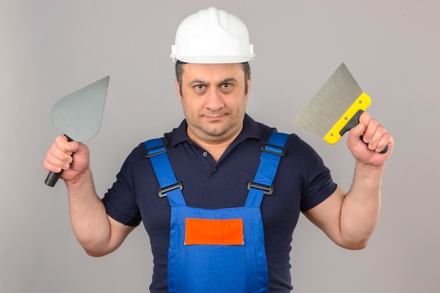 Photo gratuite builder homme portant des uniformes de construction et un casque de sécurité debout avec un couteau à mastic et une truelle dans les mains avec un visage sérieux sur un mur blanc isolé