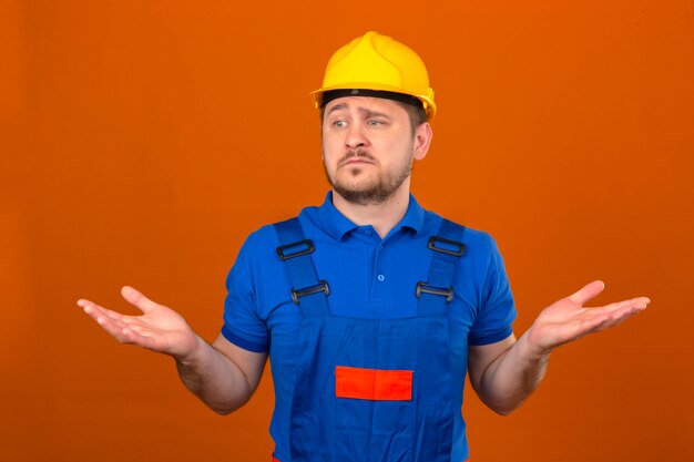 Builder homme portant des uniformes de construction et un casque de sécurité désemparé et expression confuse avec les bras et les mains soulevées concept de doute sur mur orange isolé