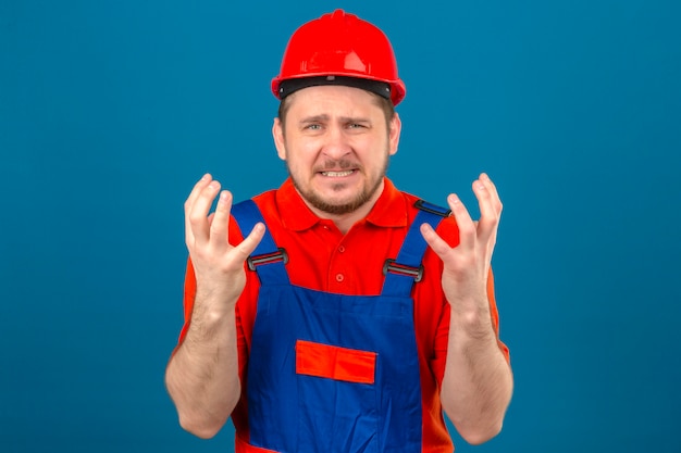Builder homme portant des uniformes de construction et un casque de sécurité fou et fou debout avec une expression agressive et les bras levés sur un mur bleu isolé