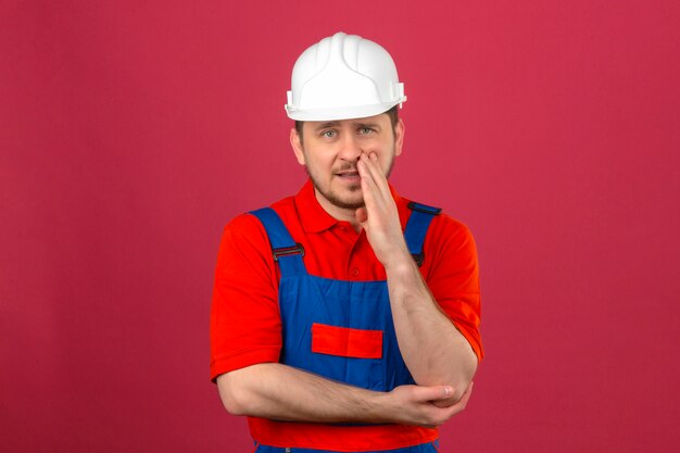 Builder homme portant des uniformes de construction et un casque de sécurité avec une main près de la bouche disant un secret debout sur un mur rose foncé isolé