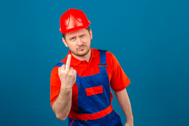 Builder homme portant des uniformes de construction et un casque de sécurité mécontent montrant le majeur à la caméra debout sur mur bleu isolé