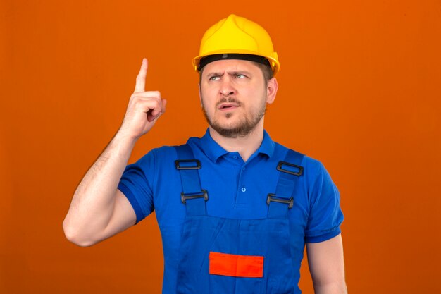 Builder homme portant des uniformes de construction et un casque de sécurité met en garde contre le danger en soulevant son doigt montrant un panneau d'avertissement avec le doigt debout sur un mur orange isolé