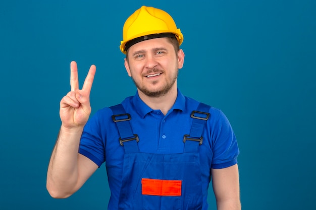 Builder homme portant des uniformes de construction et casque de sécurité montrant le numéro deux avec les doigts à côté souriant sympathique debout sur mur bleu isolé