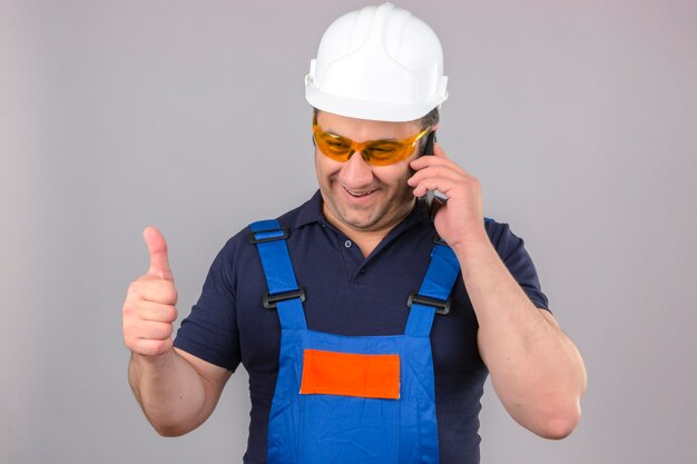 Builder homme portant des uniformes de construction et un casque de sécurité parlant au téléphone mobile heureux et souriant montrant les pouces vers le haut sur un mur blanc isolé