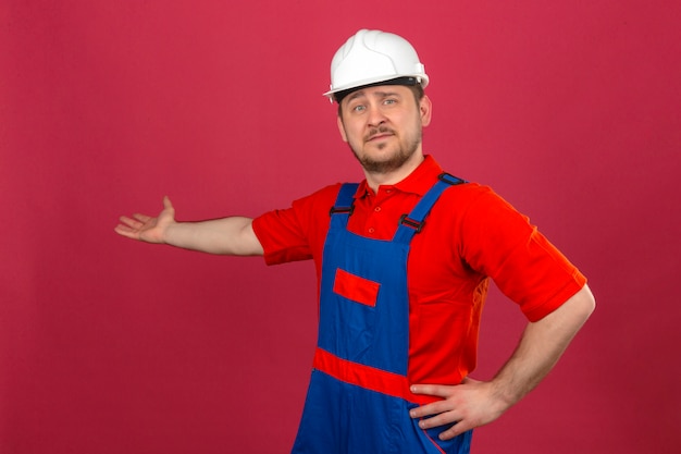 Builder homme portant des uniformes de construction et un casque de sécurité pointant de côté avec la main ouverte paume montrant copie espace souriant confiant sur mur rose isolé