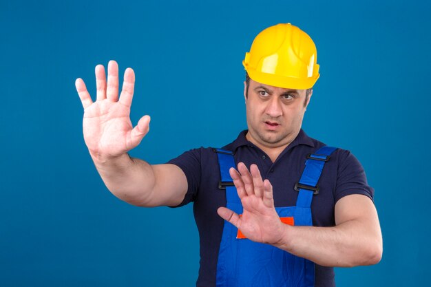 Builder homme portant des uniformes de construction et un casque de sécurité tenant ses mains en disant ne pas se rapprocher de mur bleu isolé