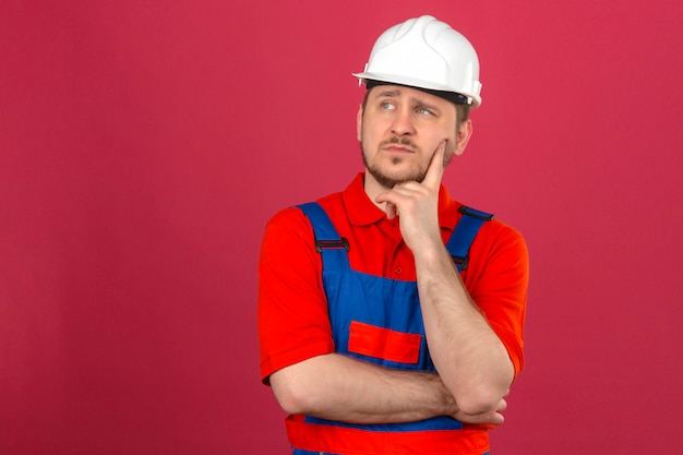 Builder homme portant des uniformes de construction et un casque de sécurité touchant sa joue et pensant regard pensif debout sur un mur rose isolé