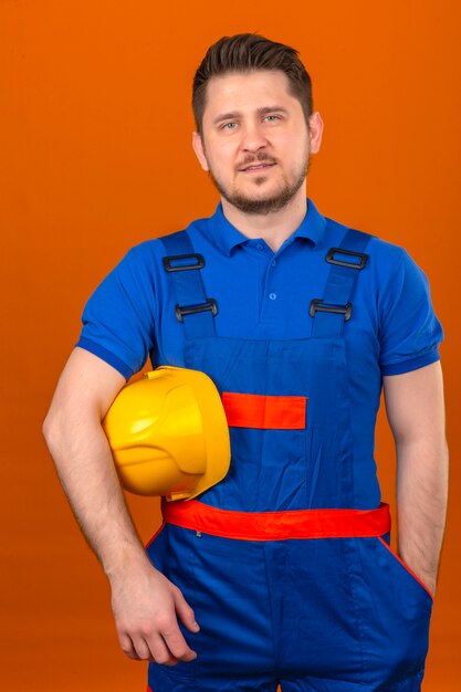 Builder homme portant des uniformes de construction debout avec un casque de sécurité à la main avec un sourire confiant sur mur orange isolé