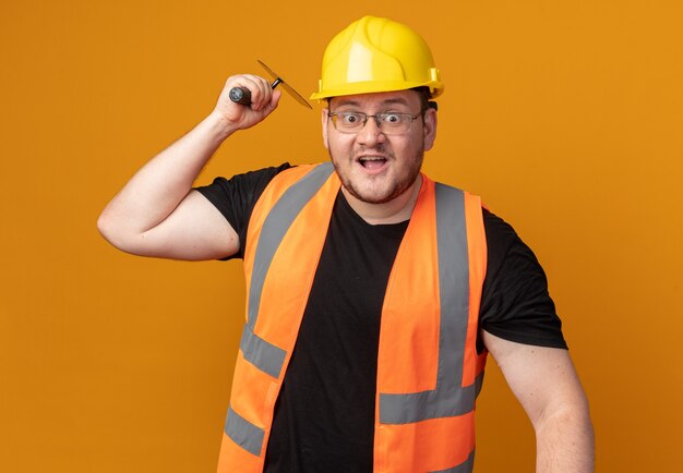 Builder man in construction gilet et casque de sécurité balançant un couteau à mastic émotionnel et surpris debout sur orange