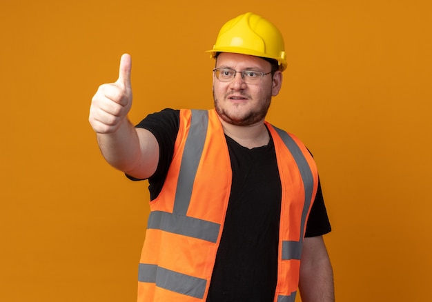 Builder man in construction gilet et casque de sécurité à côté souriant confiant montrant les pouces vers le haut debout sur fond orange