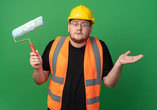 Builder man in construction gilet et casque de sécurité holding paint roller looking at camera confus haussant les épaules