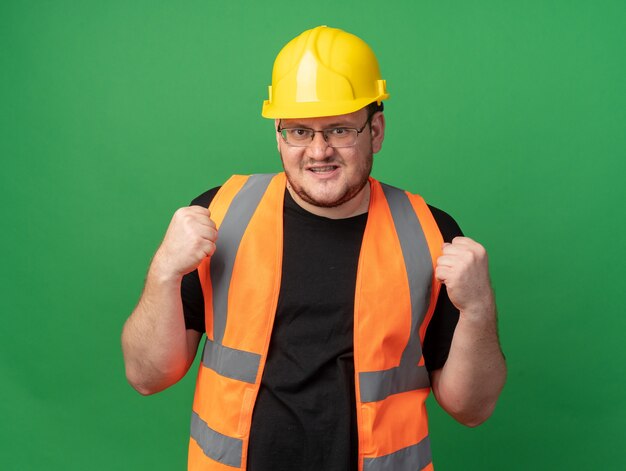 Builder man in construction gilet et casque de sécurité looking at camera smiling confiant heureux et positif debout sur fond vert