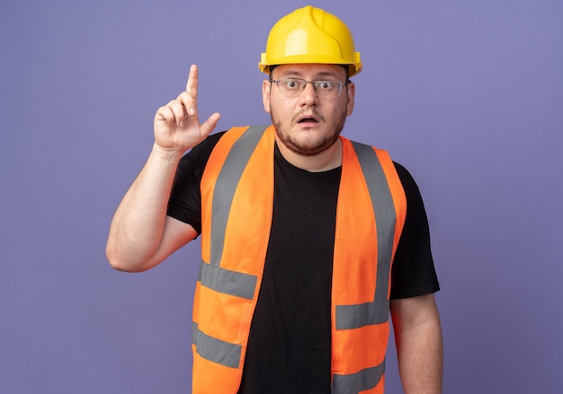 Builder man in construction gilet et casque de sécurité looking at camera surpris et inquiet montrant l'index debout sur bleu