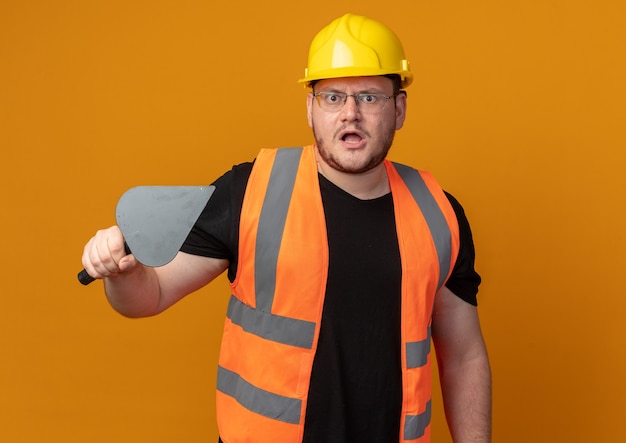 Photo gratuite builder man in construction gilet et casque de sécurité tenant un couteau à mastic regardant la caméra étant mécontent du visage en colère debout sur fond orange