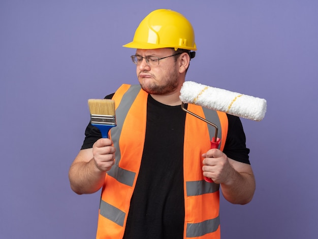 Builder man in construction gilet et casque de sécurité tenant un rouleau à peinture et un pinceau à la confusion en essayant de faire un choix debout sur bleu
