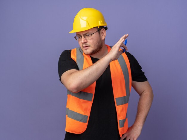 Builder man in construction gilet et casque de sécurité tenant un stylo à côté avec un visage sérieux debout sur bleu