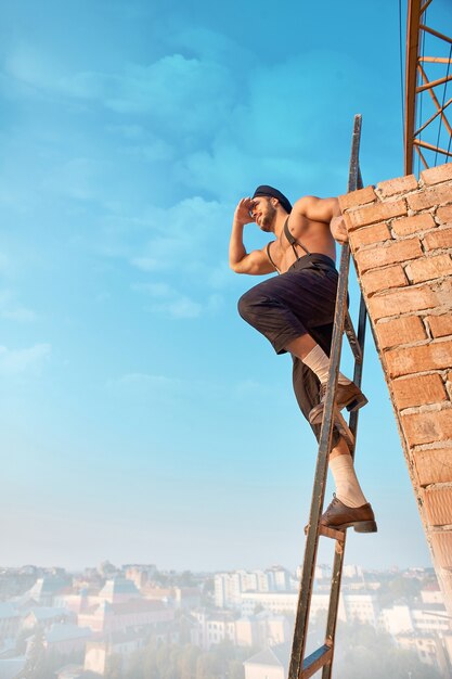 Builder s'appuyant sur un mur de briques et assis sur une échelle en hauteur. Homme au torse nu en tenue de travail tenant la main près des yeux et regardant à distance. Paysage urbain sur fond.