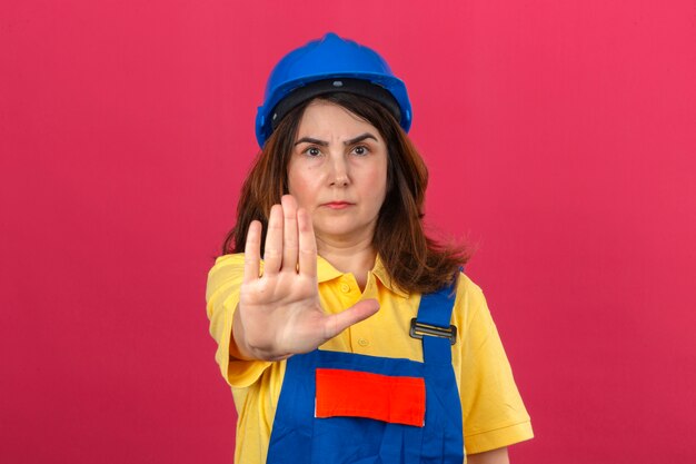 Builder woman wearing construction uniforme et casque de sécurité debout avec la main ouverte faisant panneau d'arrêt avec un geste de défense d'expression sérieuse et confiante sur mur rose isolé