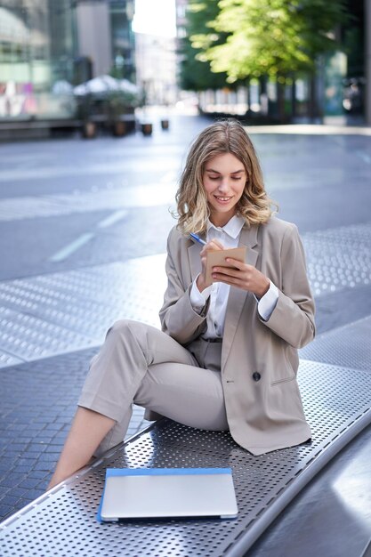 Businesswoman sitting with laptop wearing suit écrire des notes dans son planificateur d'être en centre-ville