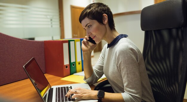 Businesswoman talking on mobile phone tout en utilisant un ordinateur portable au bureau