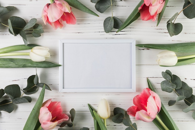 Photo gratuite cadre blanc blanc entouré de tulipes blanches et roses sur un bureau en bois