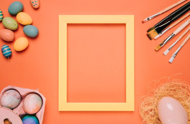 Cadre de bordure jaune entouré d&#39;oeufs de Pâques; nid et pinceaux sur fond orange