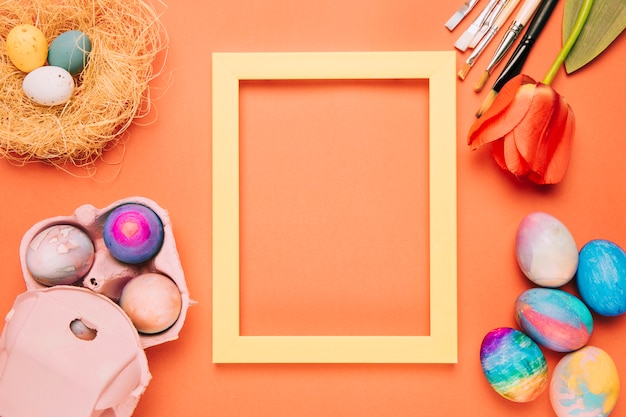 Photo gratuite un cadre de bordure jaune vide entouré d'oeufs de pâques; nid; tulipe et pinceaux sur fond orange