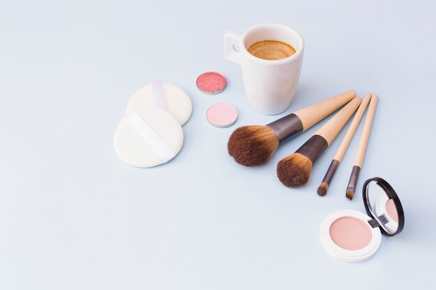 Photo gratuite café avec pinceau de maquillage; le fard à paupières; éponge et fard à joues sur fond blanc